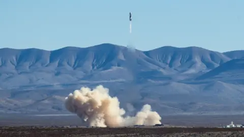 A blue origin rocket blasts off from the desert floor with mountains in the background.