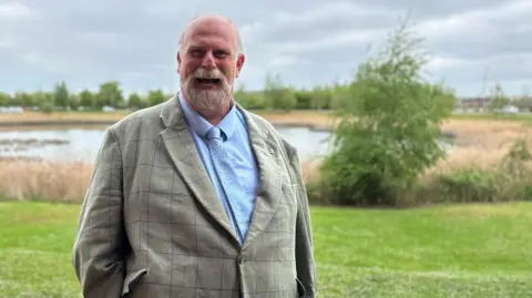 Kimberley Piper/BBC A picture of the Conservative Mayor of Bedford Tom Wootton. He is standing in front of a lake and is wearing a grey suit with black lines on it and has a light blue shirt and tie. He has a white beard and is smiling.