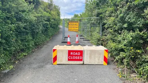A single track road flanked by trees. In the foreground is a road block and fence, warning cars the road is not available for use.