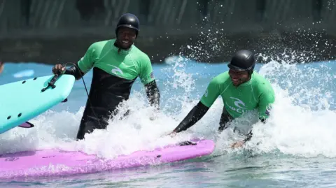 The Wave Two men stand in the water at The Wave in Bristol. They are smiling and wearing green and black wetsuits as the water foams around them