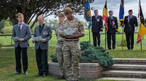 Crown Copyright Uniformed personnel and smartly dressed men with medals pinned on their jackets read from service sheets during a remembrance ceremony in a cemetery, with veterans holding flags standing behind.