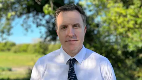 Wayne Bates looking at the camera as he stands outside. He has short dark hair, is wearing a white shirt and a striped black tie.