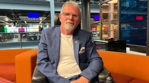 A man in a wheelchair pictured in a BBC building in Cardiff. He has short, white hair a white goatee. He is wearing a blue suit and white t-shirt and has round, clear-framed glasses.