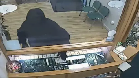 Man in hooded dark clothing reaches hand into jewellery cabinet 