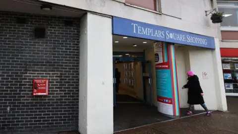 Oxford City Council A woman walking into the Templars Square Shopping Centre. The bleed kit cabinet can be seen at the side of the building on the wall.