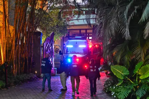 Getty Images Police HSI officers walking toward a building at night, with a police vehicle flashing lights nearby, surrounded by greenery and illuminated by various lights.