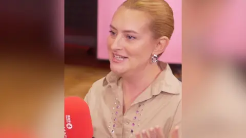 A woman with light ginger hair pulled back from her face talks to someone off-camera.