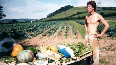 Riverford This is a photo dating back likely to the 1980s, showing Guy Singh-Watson as a young man, bare-chested, standing in a field with rows of crops. It looks like a warm summer's day. In front of Guy is a wheelbarrow with boxes of vegetables loaded on it.