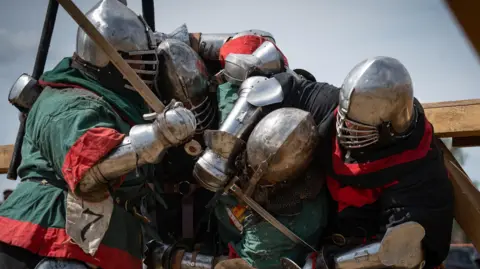 Four people wrestling in medieval-style armour and with swords in hand. Two are in green and red and another two are in red and black clothing with large metal helmets and slatted visors