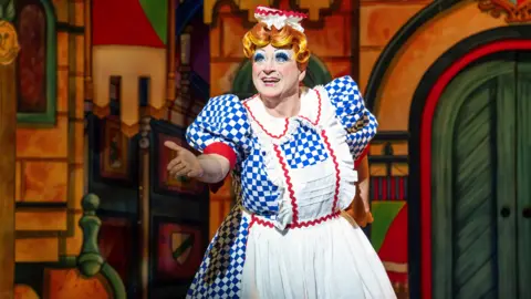 Richard Hubert Smith A scene from a pantomime shows a man in a blue and white dress, dressed as a dame, holding his arm out. There is scenery behind him. 