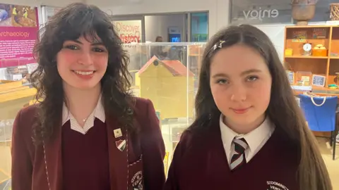Two girls in school uniform. Annabelle on the left has long curly brown hair and is wearing a blazer over a maroon jumper and red and white striped blouse. Aimee on the right is wearing a maroon v-neck sweater over a white shirt and striped black, red and silver tie