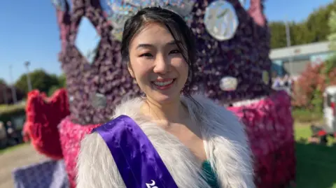 A head and shoulders shot of Erika, who wears a purple sash over a fluffy top. The main float is behind her. She has short dark hair and is smiling into the camera.