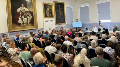 Handout Rows of people sitting in a public meeting in Great Torrington Town Hall with a man at the front of the hall addressing the crowd and old paintings on the walls