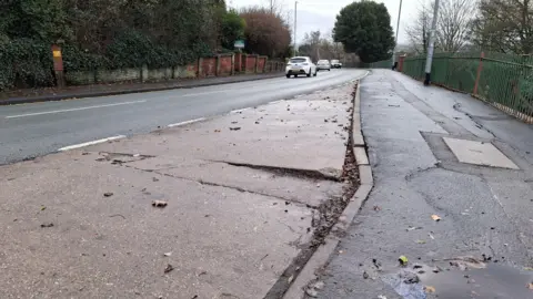 A photo of a road, concrete layby and footpath, looking downhill towards a bend. There are large cracks in the layby and smaller cracks on the footpath. To the left of the road, a weathered brick wall stands in front of thick green foliage. To the right, green railings stand above an embankment, with bare trees rising behind it. Cars can be seen in the middle distance.