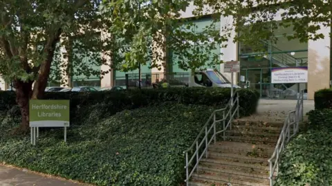 Google Exterior of a modern library building with large glass windows and an entrance reached by steps and metal railings. A sign in the foreground reads “Hertfordshire Libraries,” with another sign by the entrance for “Central Stacks & Performing Arts Service.” Trees and ivy-covered banks frame the pathway and steps leading up to the building.