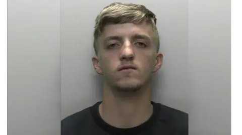 A mugshot of Owen Dunstan. He is staring at the camera and has short blond hair and is wearing a black top.