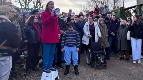 Fleur Anderson stands on a small step stool using a megaphone to address a crowd gathered at the entrance to Hammersmith Bridge, with people of all ages listening and a police officer standing on the bridge above.