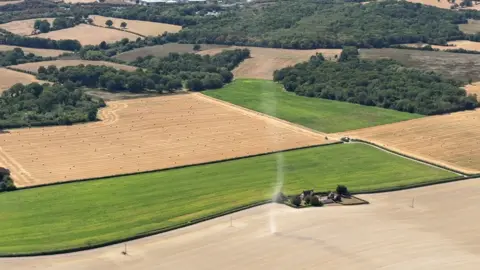 A dust devil crosses a dusty field, as seen from the air. The twister is passing a farmhouse.