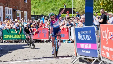 Phil Crow Lauren Dickson, a female cyclist, riding across a white finish line on a cobbled street. She is wearing blue, pink and white cycling kit and is riding on a light blue bike. A cyclist in black kit, on a black bike, is approaching the finish line behind her. Barriers with adverts line line the route, with groups of spectators watching from behind them. 