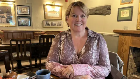 Sarah Stenton has light brown hair and is sitting in a pub at a table. She is wearing a pink and white patterned blouse, and is smiling at the camera.