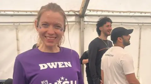 Ceiridwen after she completed her race standing proudly in a tent. Her blonde hair is tied back and she is smiling. She is wearing a purple My Wish t-shirt with her race number on the front