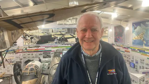 Photograph of Terry Barnes, the Chairman of the Avro Heritage Museum in Stockport. He is pictured on a balcony overlooking the inside of the museum.