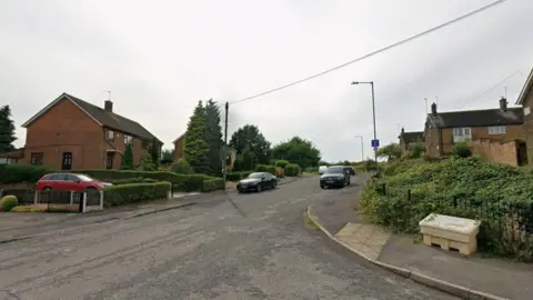 Google A Google streetview of a cul-de-sac with cars parked on both sides and houses. 