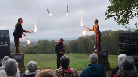 Two fire jugglers on plinths throwing flaming batons to each other in front of a small crowd of people