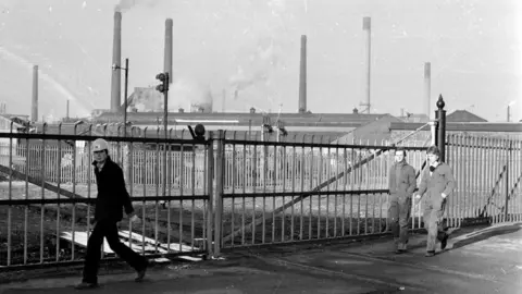 Getty Images A black and white photo of Corby's steelworks. It is a huge factory with several chimneys. It is in the distance behind a fence. A man in a hard hat looks at the camera as he walks in front of the fence. Two other men in boiler suits are walking past a short distance behind him.