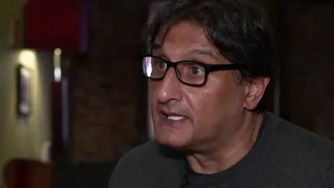 A head-and-shoulders picture of Ishy Din. He has short black hair, dark eyes and wears glasses with a black rectangular frame. He is wearing a grey fine-knit jumper and is looking out of shot to a reporter interviewing him. The background is blurred.