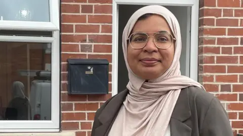 A woman wearing glasses and a headscarf stands outside a house smiling for the camera.