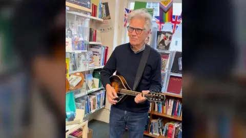 Oxfam A man in a grey jumper and glasses plays a black and orange mandolin in front of a book case.