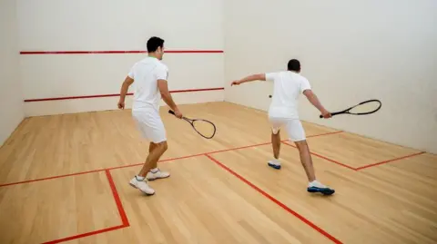 Getty Images Two men playing squash in a sports hall, wearing white shorts and T-shirts and trainers.