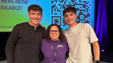 Jordan and Cian Adams stand with their arms around BBC Hereford and Worcester presenter Tammy Gooding. Jordan is wearing a long-sleeved dark grey t-shirt, Cian is wearing a light grey short-sleeved t-shirt and Gooding has a purple BBC Hereford & Worcester t-shirt on. 