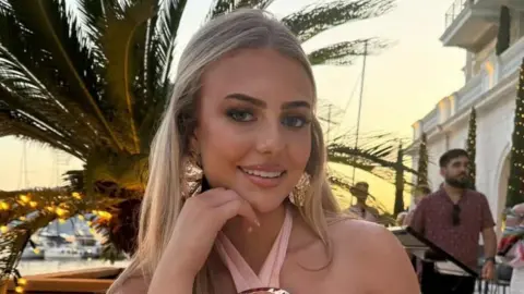 Daisy Willis Daisy is sitting down with a palm tree in the background. Daisy has blonde hair- worn half up half down. She has a pink halter neck top and big gold earrings.