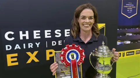 Lowri Wright holds a trophy in one hand and a rosette and certificate in the other. She has long brown hair and is standing in front of signage saying Cheese Dairy Expo