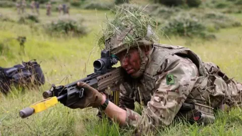 MOD A soldier on the ground in long grass pointing a long gun ahead of him during a military exercise.