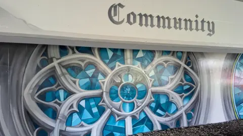 Wychavon District Council A large blue and silver mural depicting a round shape with a floral pattern in the middle. Above the painting the word "community" has been painted in old-fashioned font.