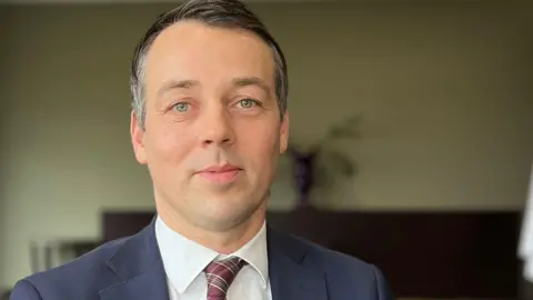 BBC Filip Hermann is sitting in a hotel conference room. He is wearing a navy suit and red, checked tie and is looking at the camera.