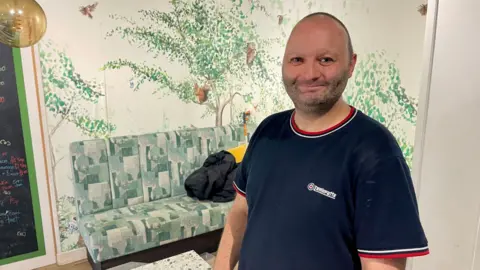 A man with a shaved head and a navy T-shirt with red and white trim is standing in a room with a painted tree on the wall.