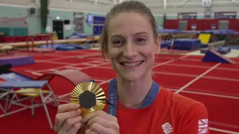 A smiling Bryony Page posing with her Paris 2024 Olympic gold medal. She has fair hair which is tied back. She is wearing a red Team GB top and is standing in a large sports hall with a red floor. Behind her are a number of blue trampolines and ramps.