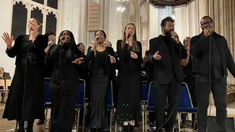 Jon Wright/BBC Six people stand, dressed in black, singing into microphones inside a cathedral 