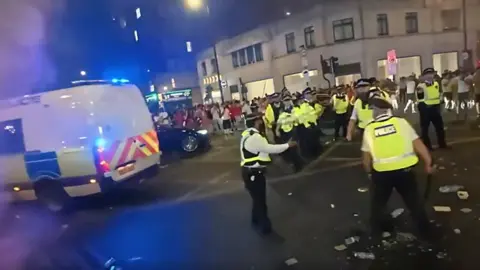 Police officers stand in the street, a police van behind them, and crowds in the background.