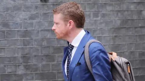 Morgan McSweeney wearing a dark blue suit, with a rucksack on his back, walking against a building with grey bricks 