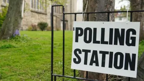 A general view during local elections outside a polling station on May 05, 2022 in Sunderland, England. A black and white sign reading: "Polling station" is tied to a black metal gate. there is a grassy field behind it that leads to a tall stone church.