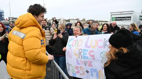 Greg James smiling at the crowd, two women are holding a sign that reads "Good luck Greg, love Jess + Ma.." . He's a white man with curly brunette hair. He's wearing a bright yellow puffer coat. There are crowds of people at the metal barrier he's smiling at them.
