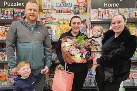 Supplied The group is standing in front of a magazine rack in the store and smiling brightly at the camera. On the right, Elisha Mcgeehin is holding toddler Blake. Molly Haggath is standing in the middle, holding a large bouquet of pink flowers and a gift bag. Mcgeehin's partner and other son are standing on the left.