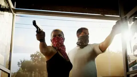 Rockstar Games Screenshot shows a male and a female character bursting through the glass front doors of a convenience store. Both have their faces covered by bandanas and each points a pistol into the premises. Behind them the late afternoon sun lights up the background, creating a hazy effect.