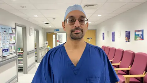 Saurav stands in a hospital waiting area, wearing a royal blue scrub top and a light blue scrub cap. He's also wearing a pair of dark-framed glasses. The hallway has white walls, floor, and ceiling, and has the bright fluorescent lighting typical of a hospital. Over Saurav's left shoulder, there is a row of five waiting room chairs, and over his right, are two side rooms.