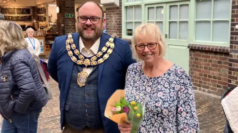 Mayor councillor Martin Williams wearing his traditional mayoral neck piece stood next to Lorraine who is holding a bunch of colourful flowers.